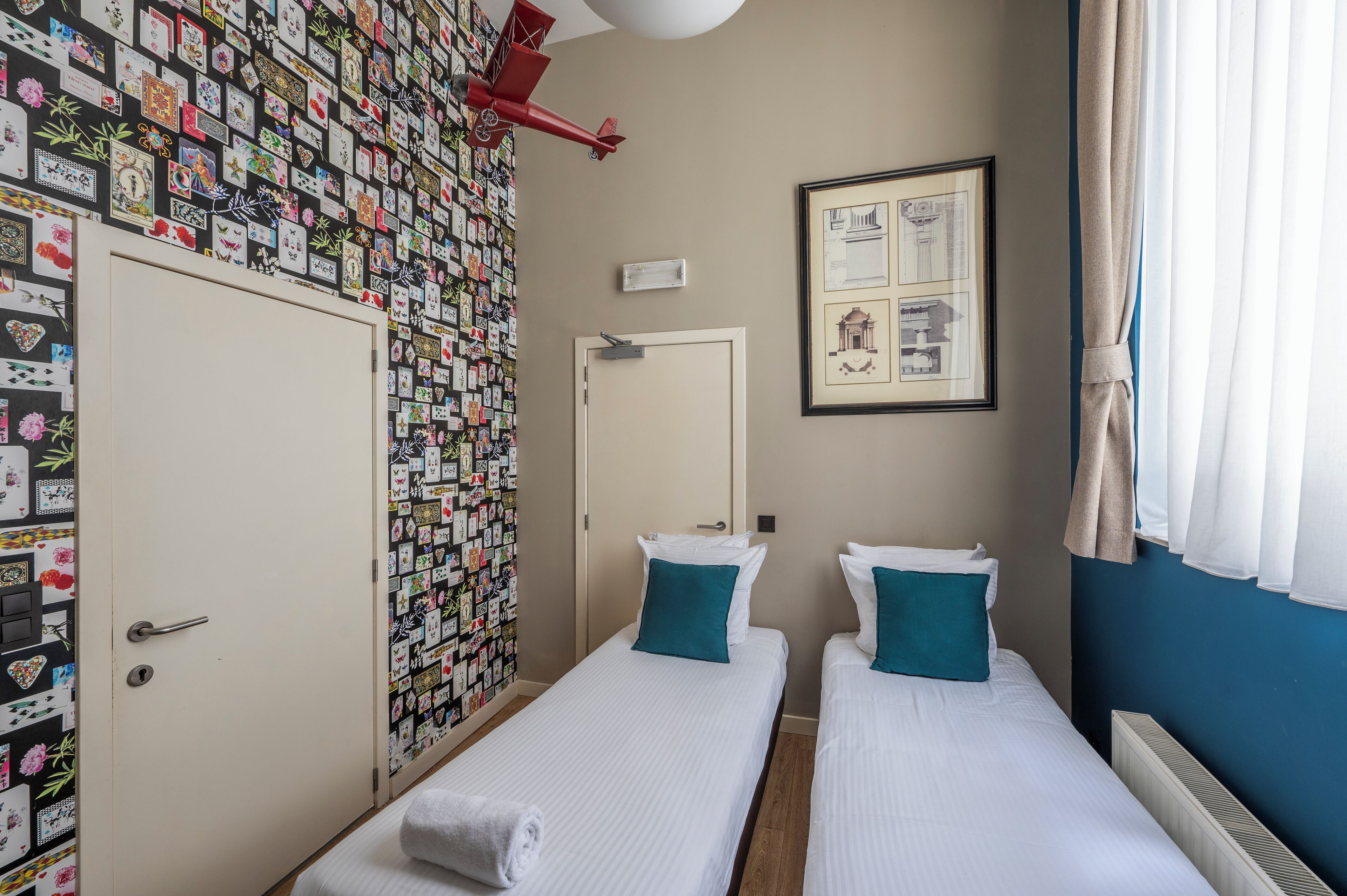 Una habitación con dos camas individuales, un armario, una pared decorada con postales y un dibujo arquitectónico enmarcado.