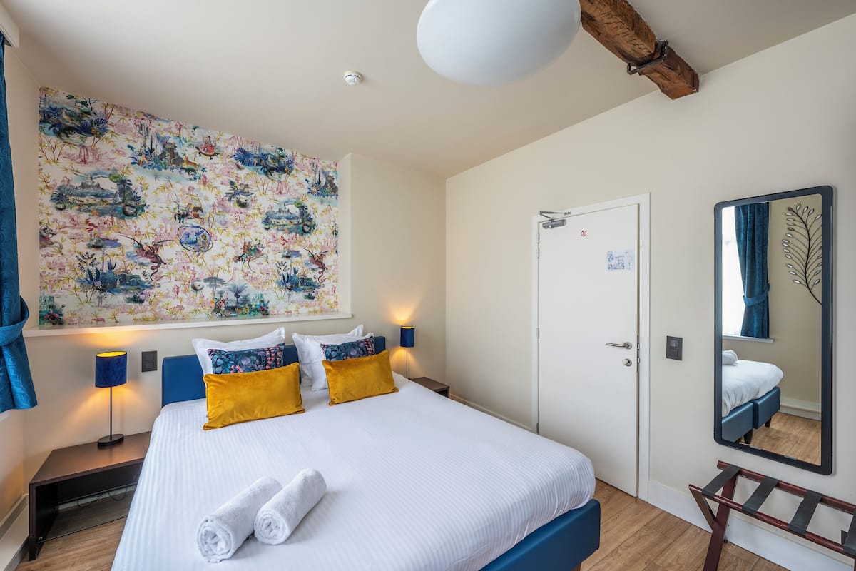Habitación de hotel con una cama grande, una mesita de noche, una lámpara, un cuadro en la pared, una puerta y un espejo.