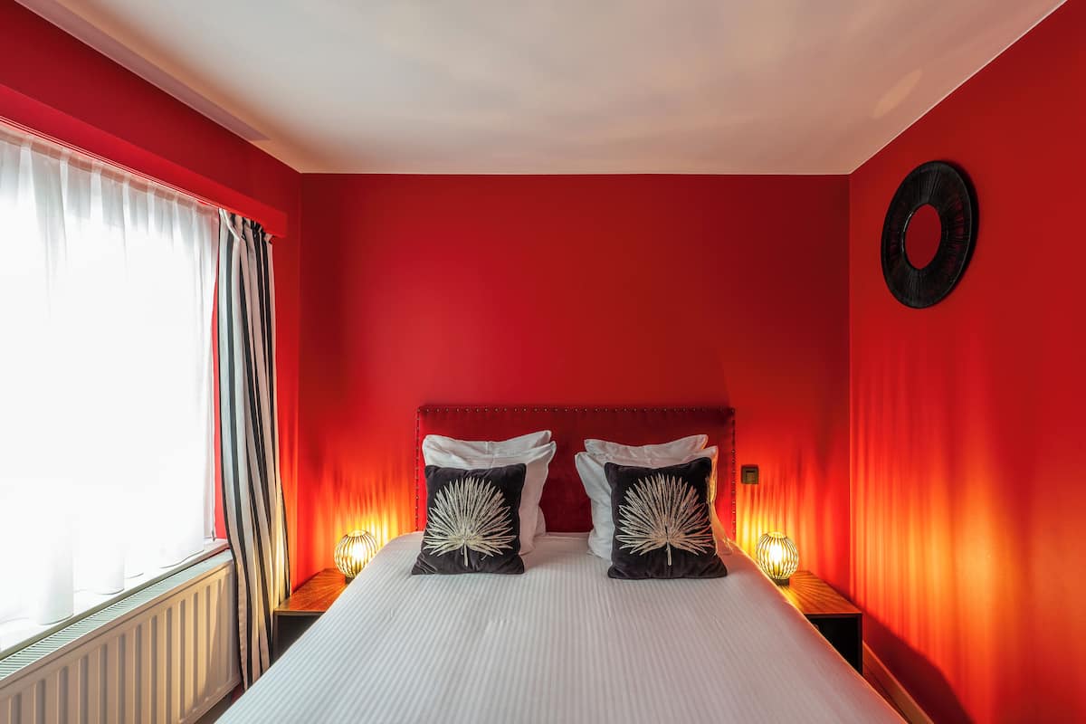 Una habitación con pared roja, cama con ropa blanca, dos almohadas negras con diseño de hojas, un adorno negro redondo en la pared y una ventana con cortinas.