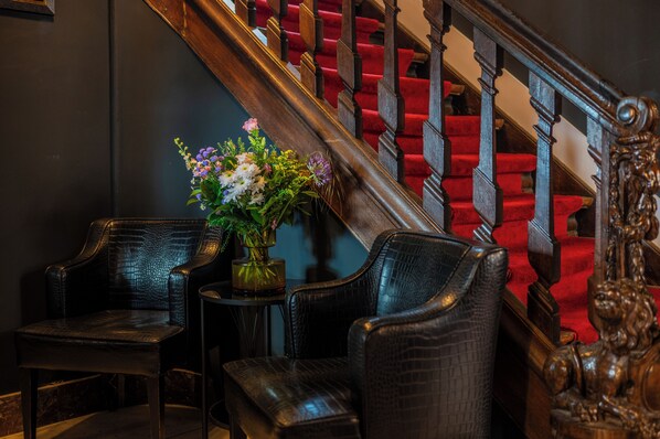 Una escalera con peldaños alfombrados en rojo y barandillas de madera, flanqueada por dos sillones de cuero y una pequeña mesa con un jarrón de flores.