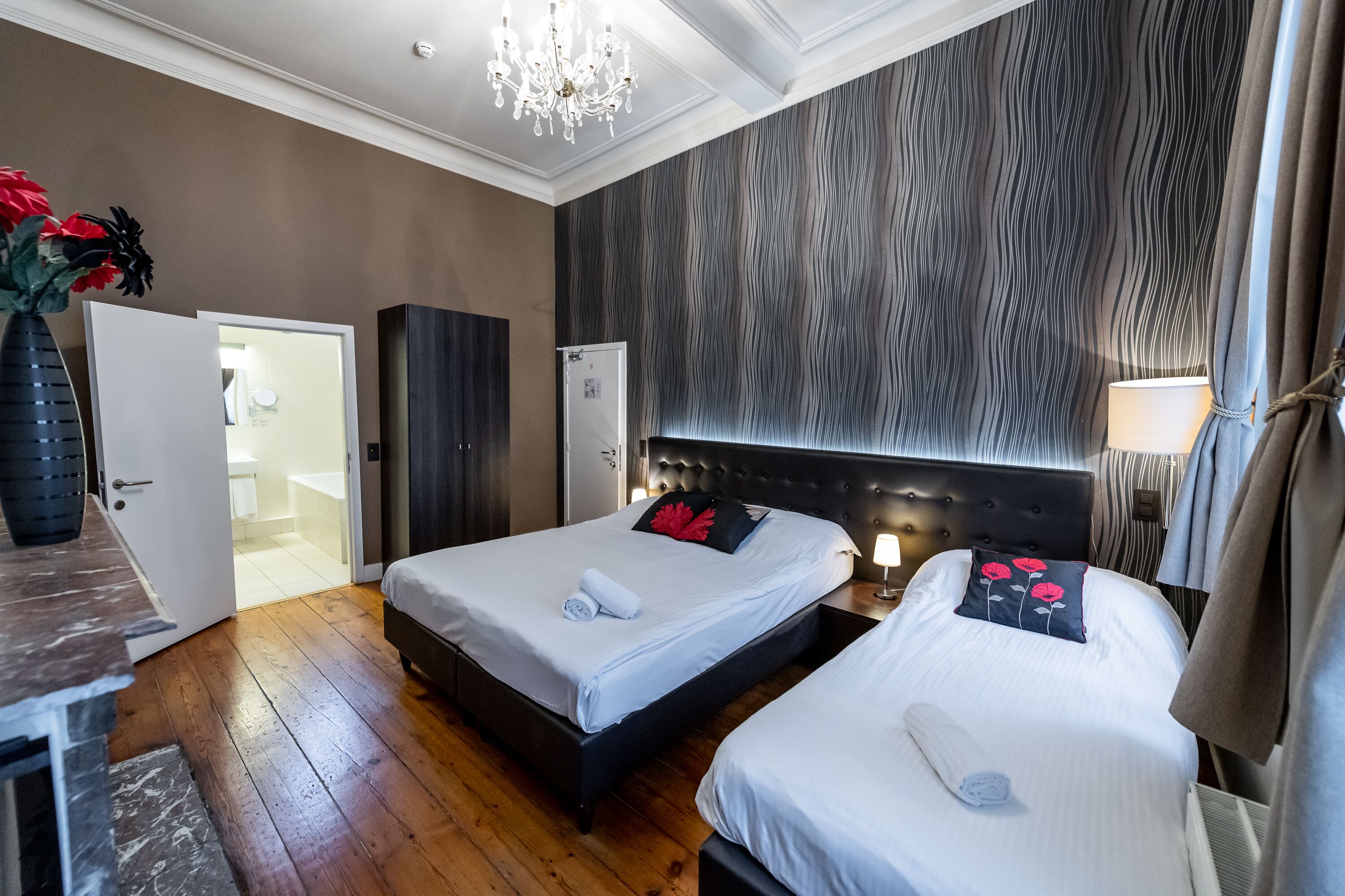 A hotel room with two beds, a chandelier, a wardrobe, and a bathroom visible through an open door.