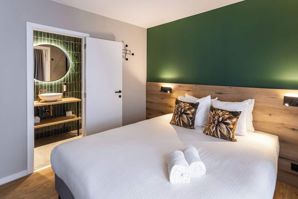 Una habitación de hotel moderna con una cama grande, cabecera de madera, pared de acento verde, espejo redondo y lavamanos empotrado con repisa de madera.