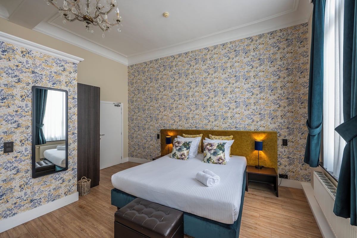 Una habitación de hotel con una cama grande, una banca, un espejo y una araña.