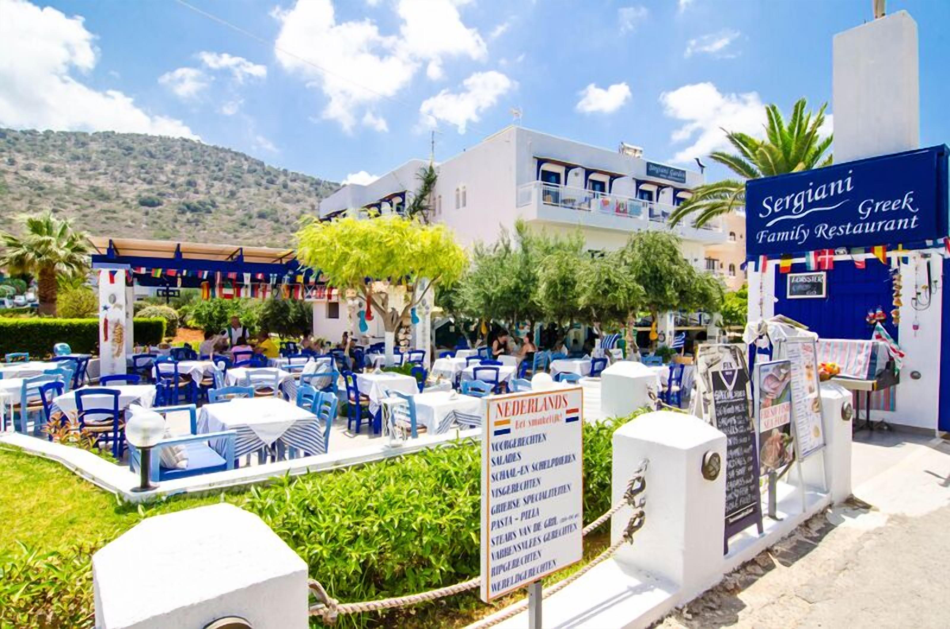 Een terras met blauwe stoelen en witte tafels, een bord met de tekst Sergiani Greek Family Restaurant, en een menubord waarop verschillende gerechten staan.