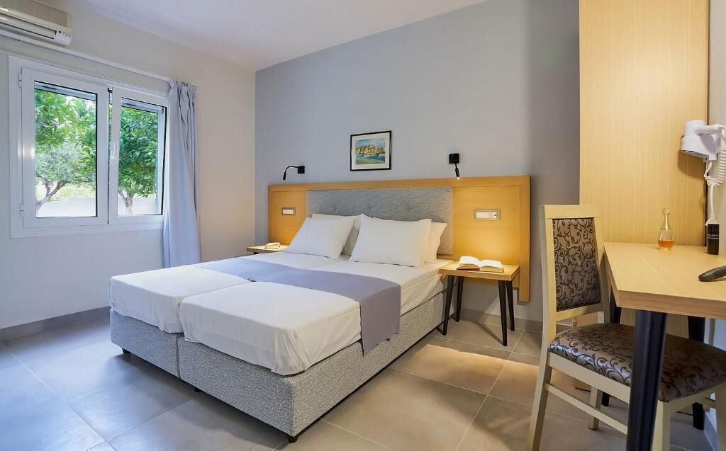 A hotel room with a double bed, a desk, a chair, a nightstand, and a window with curtains.
