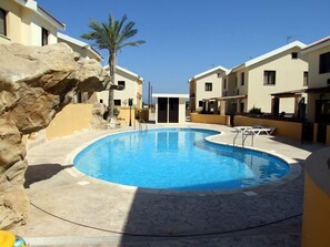 A swimming pool surrounded by buildings with a stone wall and palm trees. - Pyla View Villas, Larnaca (Livadia)