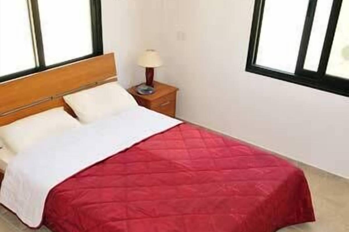 A neatly made bed with a red quilt, a wooden headboard, and a bedside lamp on a nightstand.