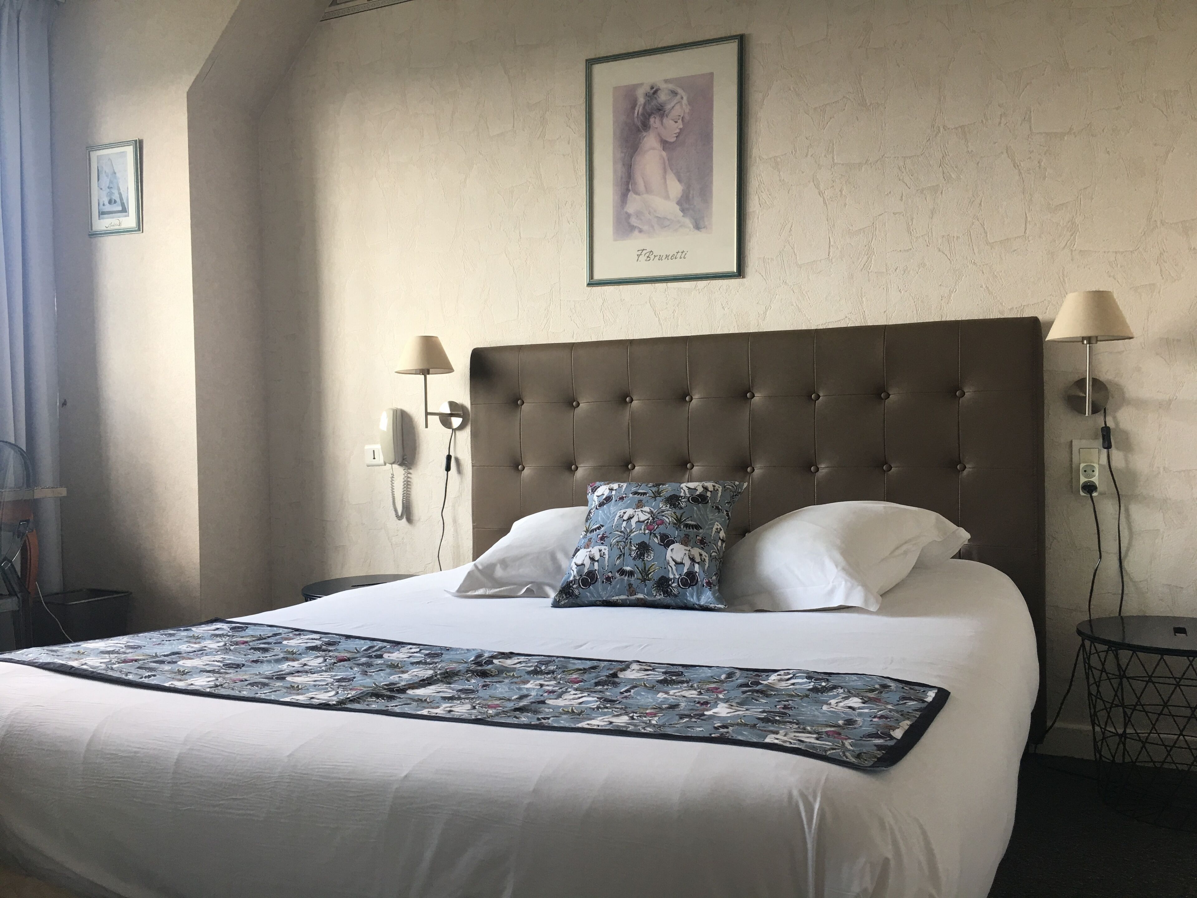 Een netjes opgemaakt bed met een gebloemde plaid, een gestoffeerd hoofdbord en een ingelijst schilderij aan de muur.