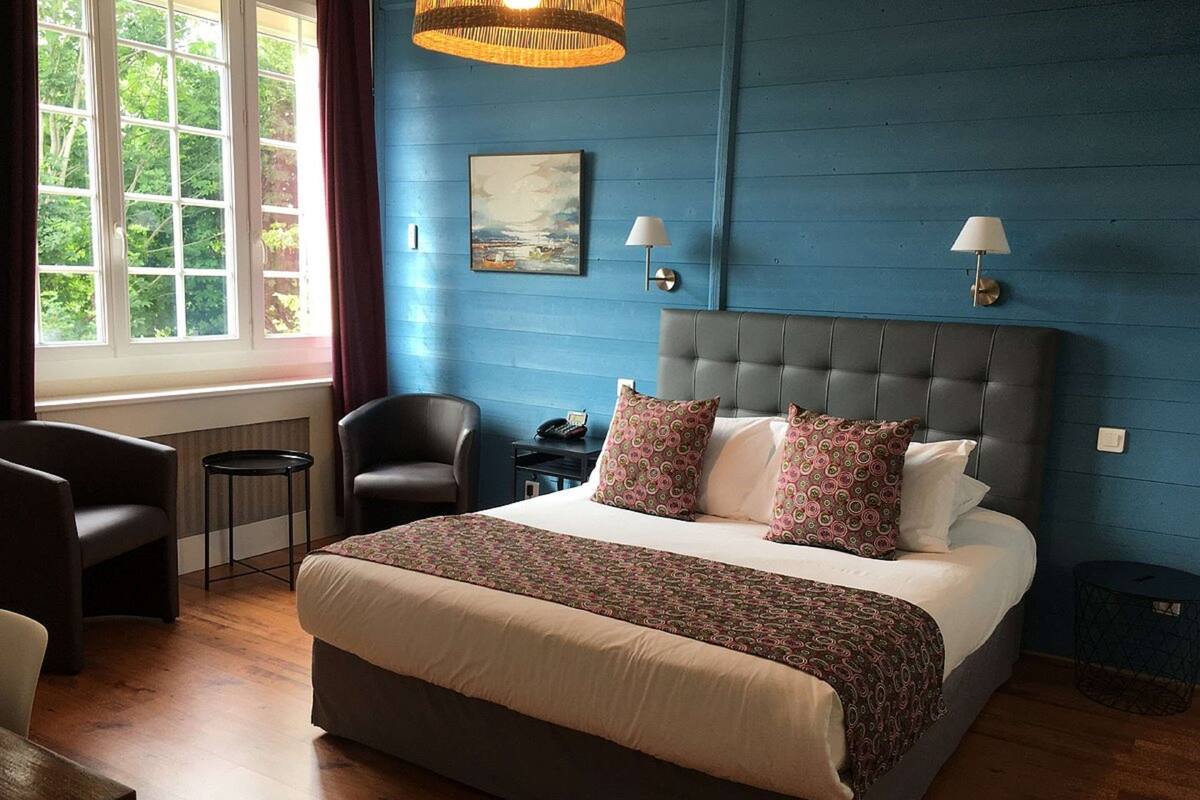 A bedroom with a large bed, two bedside lamps, a chair, a small table, and a window with curtains.
