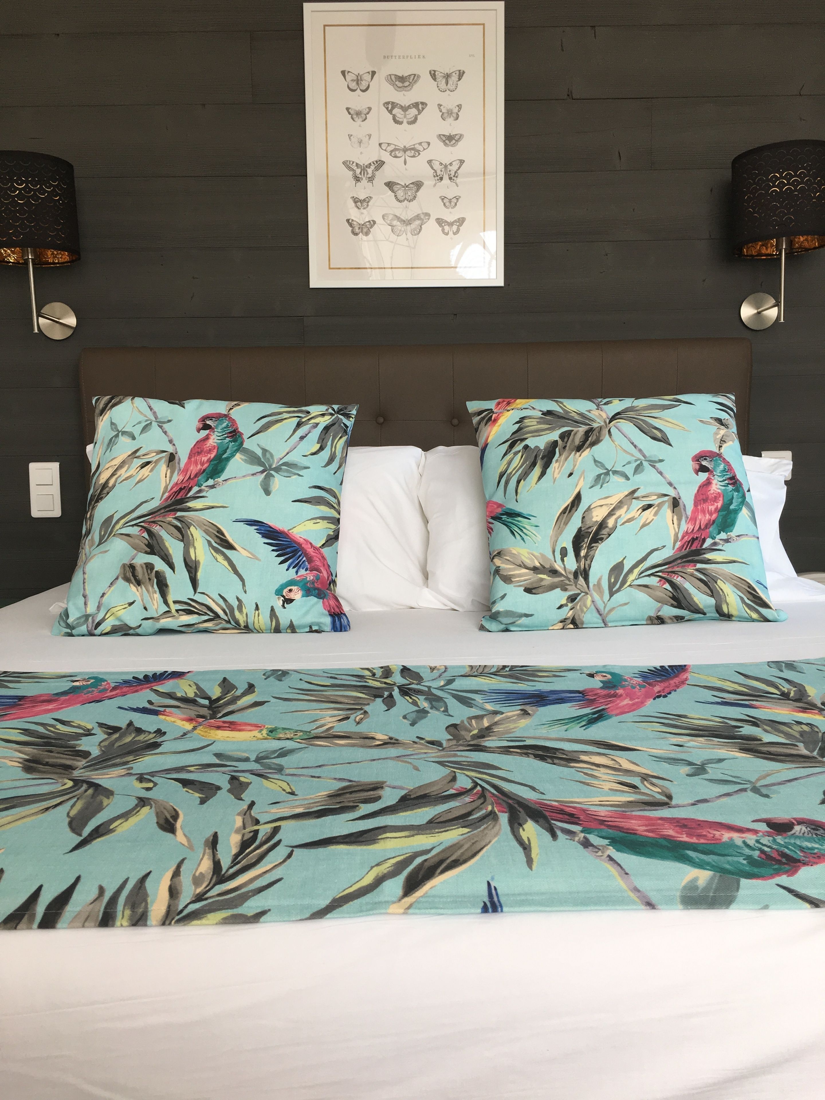 Un llit amb roba de llit amb motius florals i tropicals, una obra d'art d'una papallona emmarcada a la paret i dues làmpades penjades a la paret.