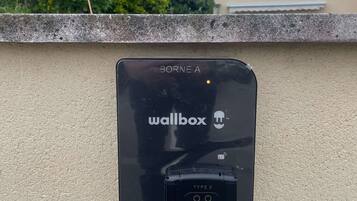 Caja de carga mural etiquetada como "BORNE A" y "wallbox" con puerto de carga "TYPE 2", sobre un muro con textura y un seto verde, con un edificio al fondo.