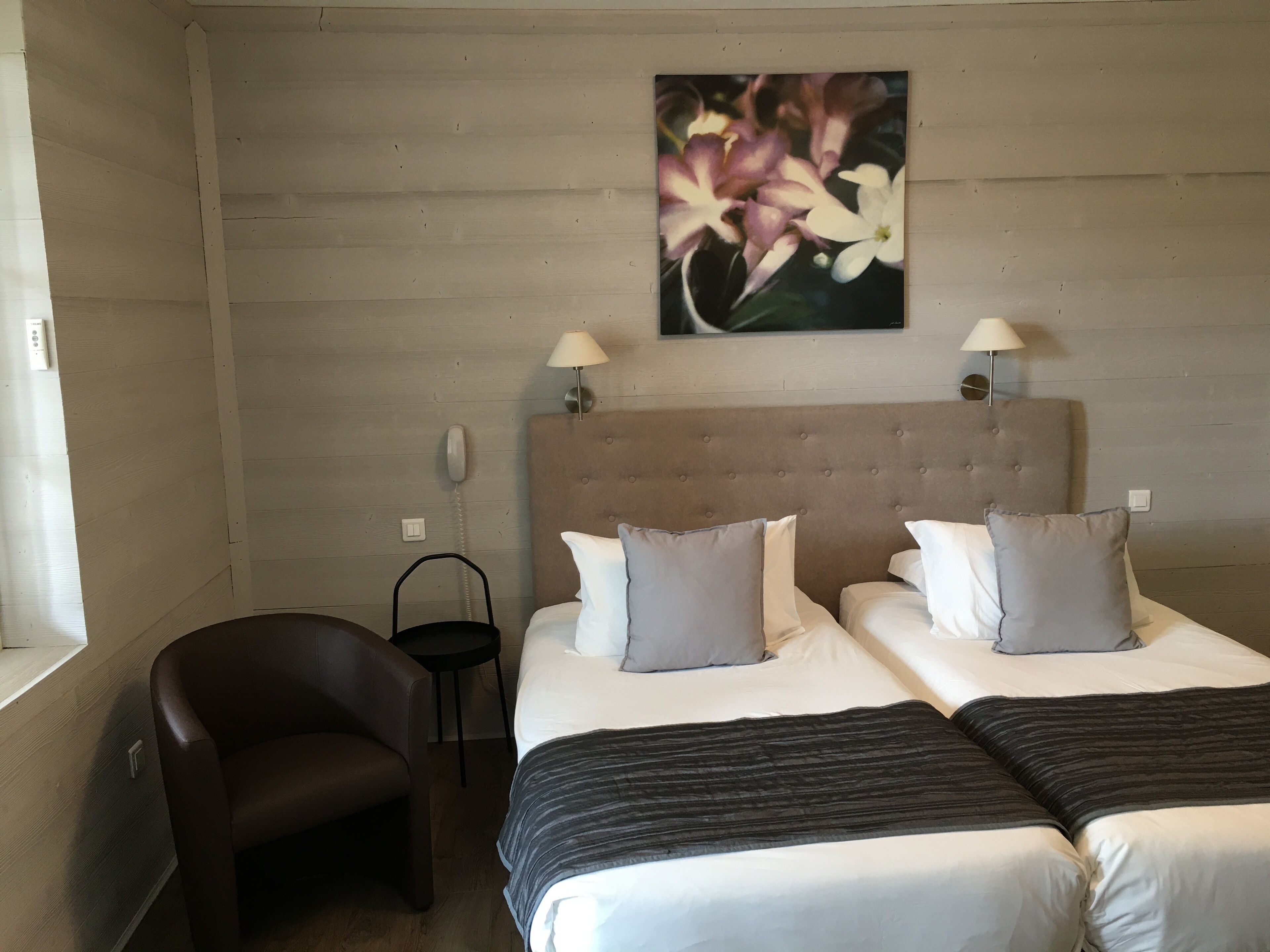 A hotel room with two beds, a chair, a bedside table, a lamp, and a painting on the wall.