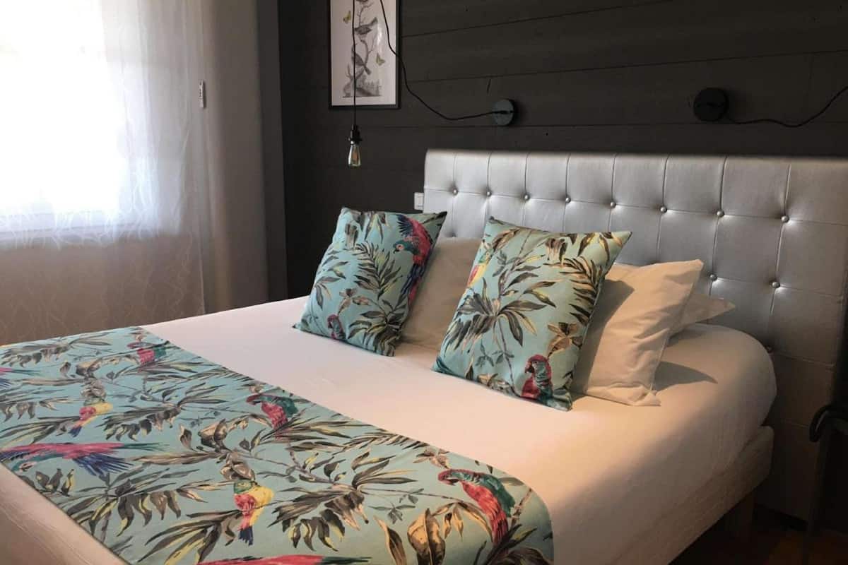 A bed with a floral bedspread, two pillows with tropical prints, a tufted headboard, and a wall-mounted artwork.
