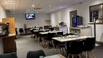 A conference room with multiple tables and chairs, equipped with a large screen for presentations, a kitchenette, and a coffee machine.