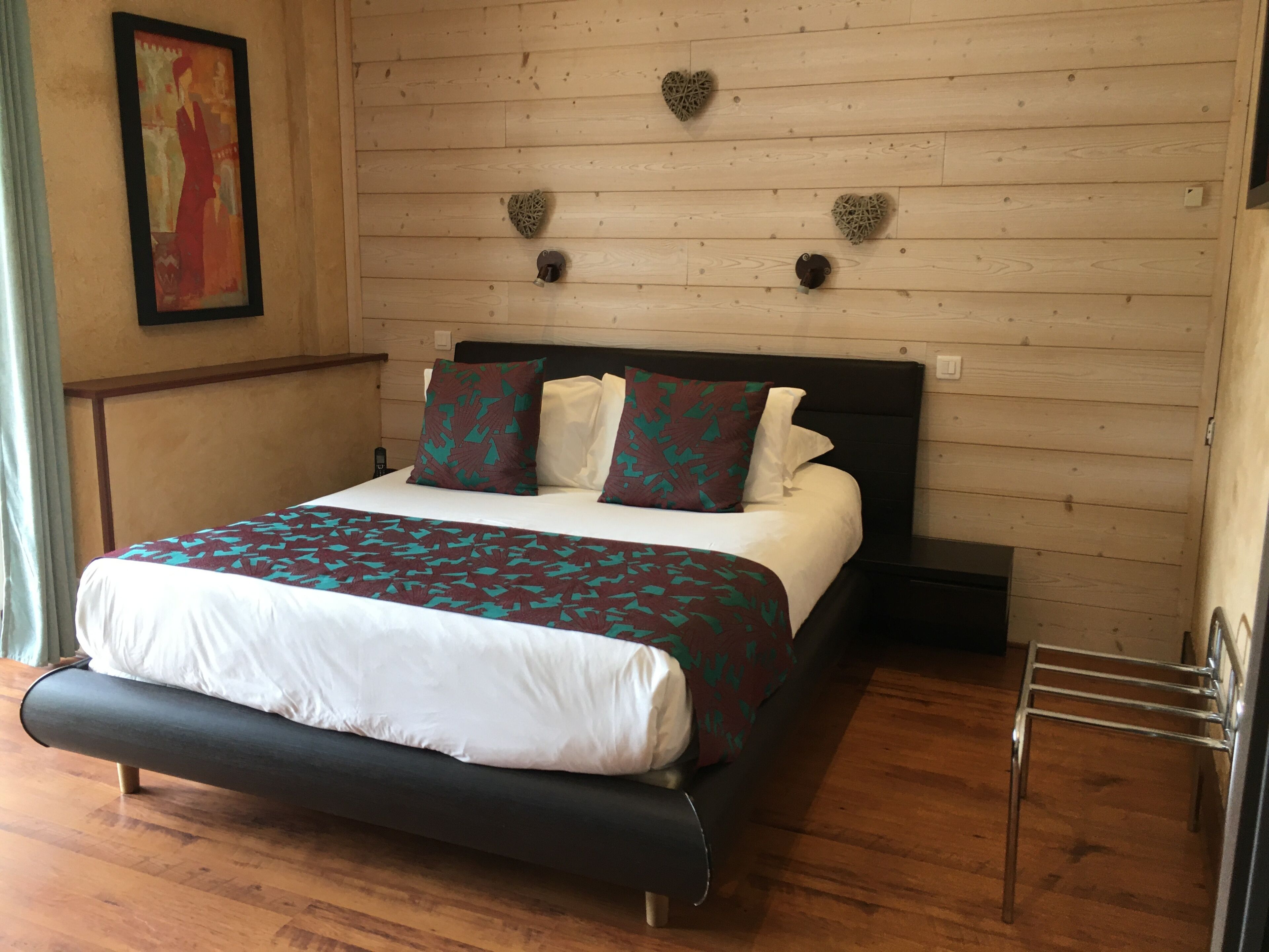 Een net opgemaakt bed met een beddegoed met patroon en kussens, een houten hoofdbord, hartvormige decoraties aan de muur, en een houten vloer.