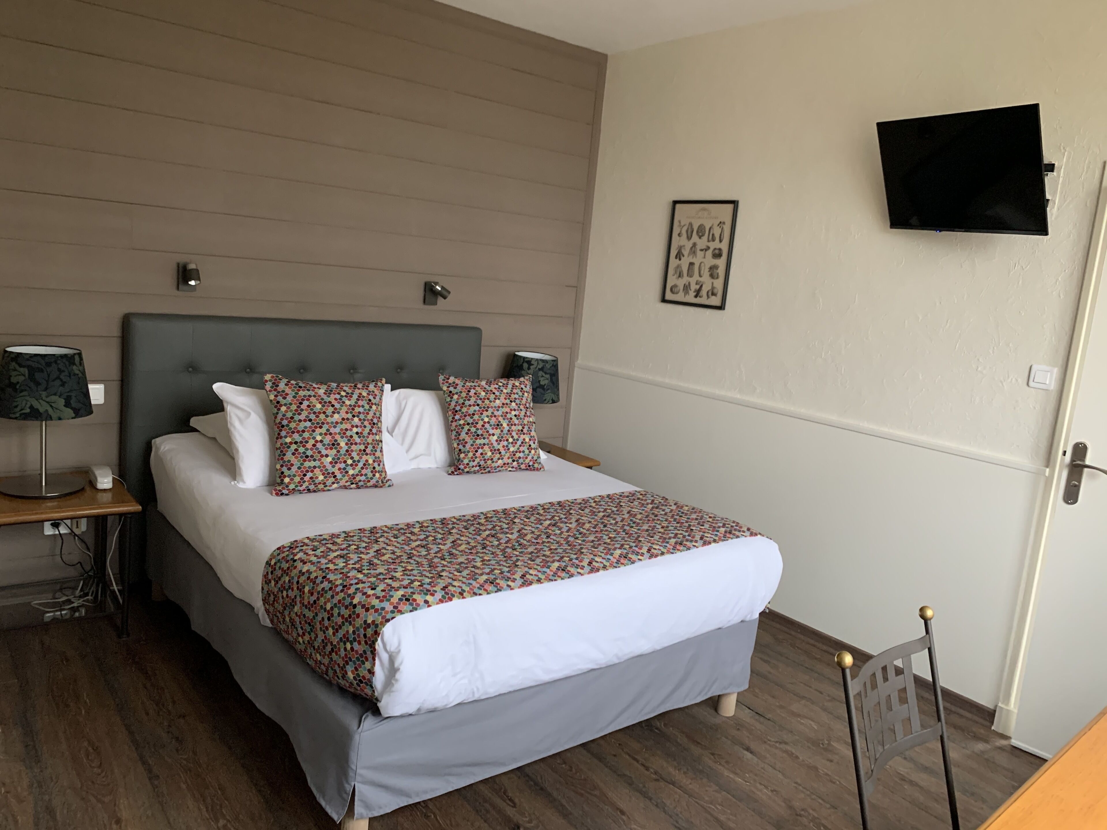 Habitación de hotel con cama, televisión montada en la pared y mesita de noche con lámpara.