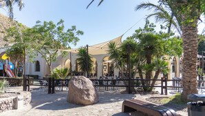 Un espacio al aire libre con pavimento de piedra, una roca grande, un banco y una zona de estar con sombra, mesas y sillas.