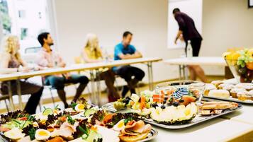 Un salón de conferencias con asistentes sentados en mesas, una pantalla de presentación y un bufé con diversas opciones de comida.