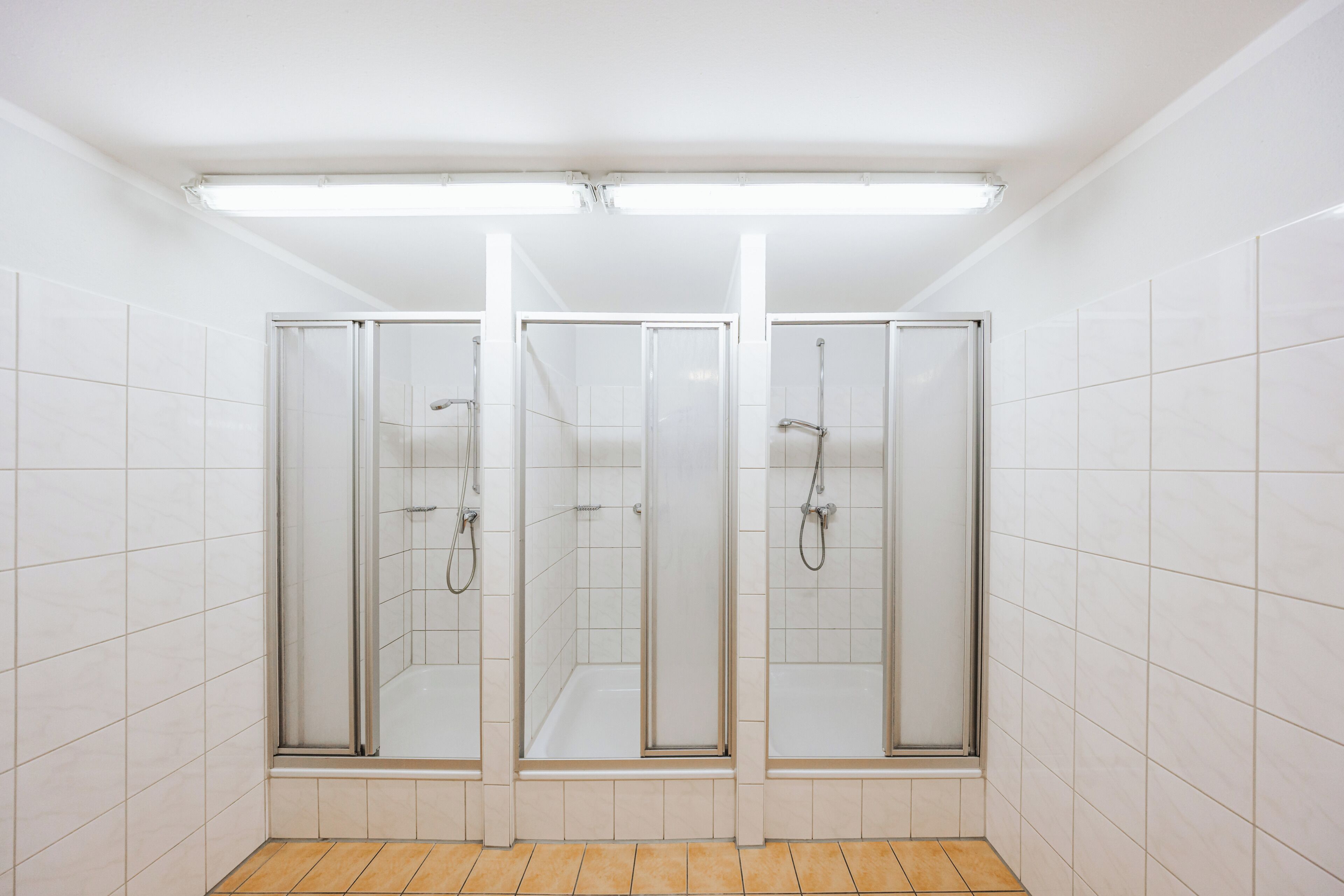 A bathroom with three glass-enclosed shower stalls, white tiled walls, and a wooden floor.