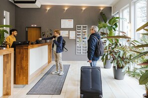 A reception desk with a wooden counter, a sign above it, and a digital screen. There are potted plants and a large window in the background.