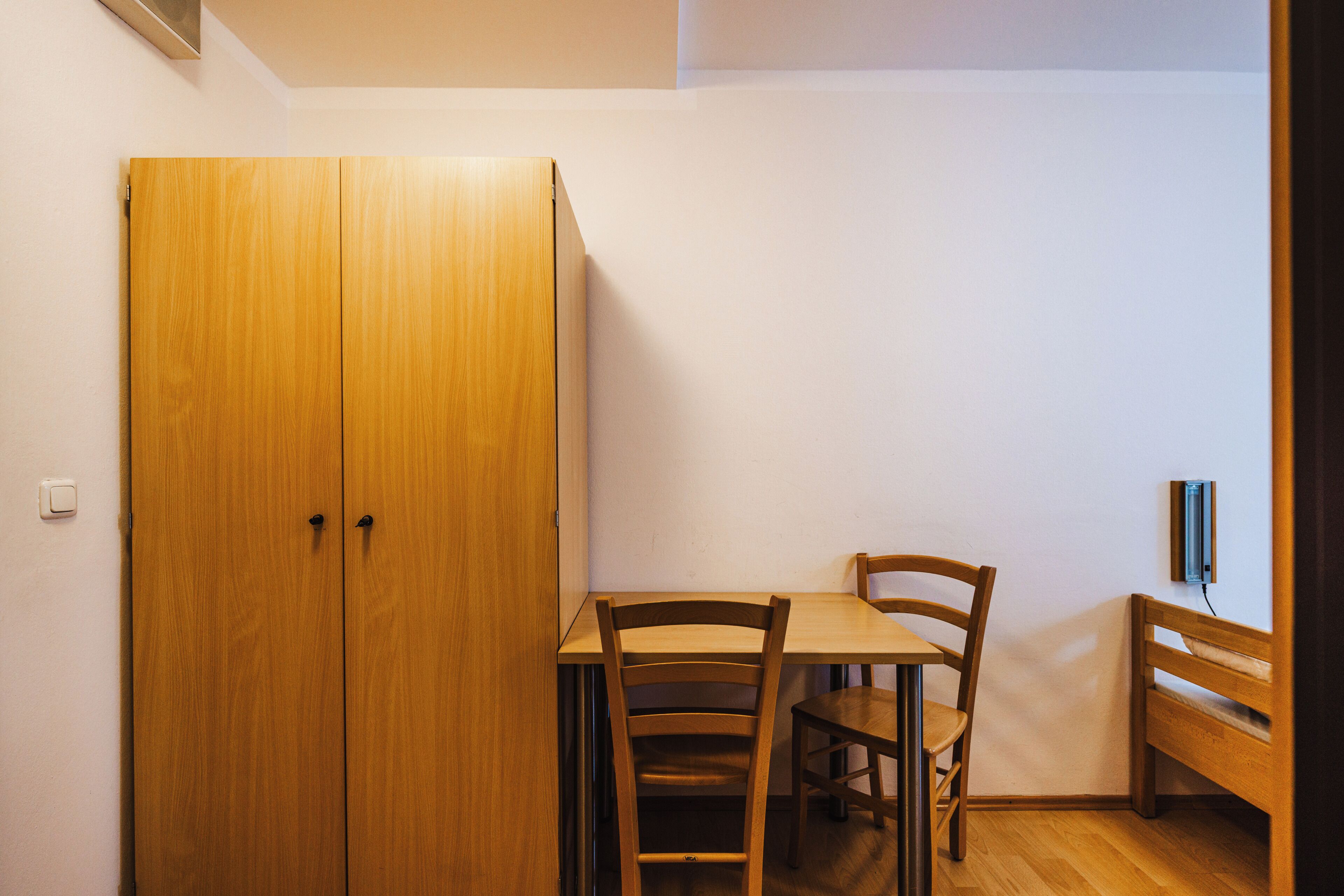 A room with a wooden wardrobe, a table with two chairs, and a bench.
