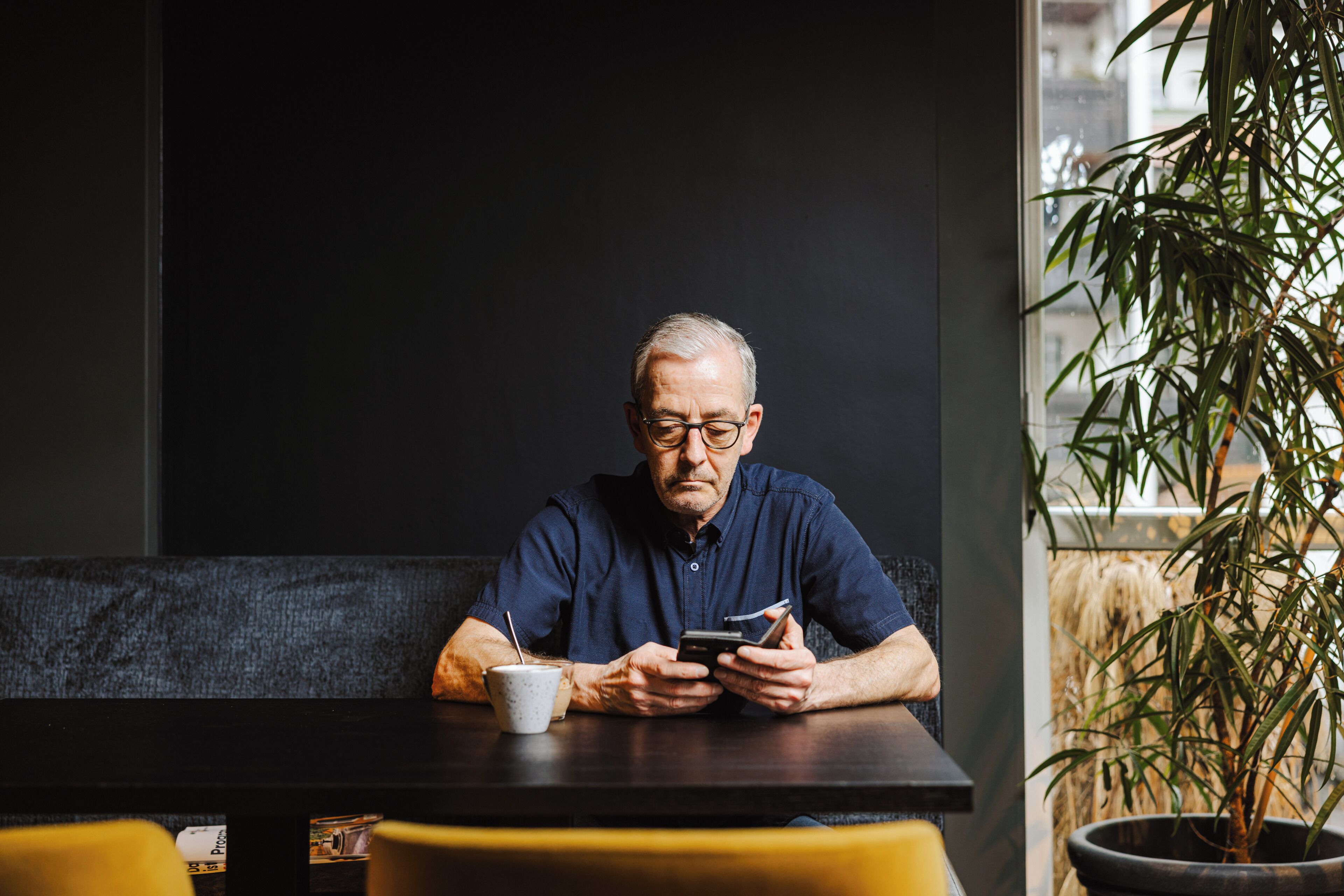 A man sitting at a table with a smartphone, a cup, and a plant in the background.