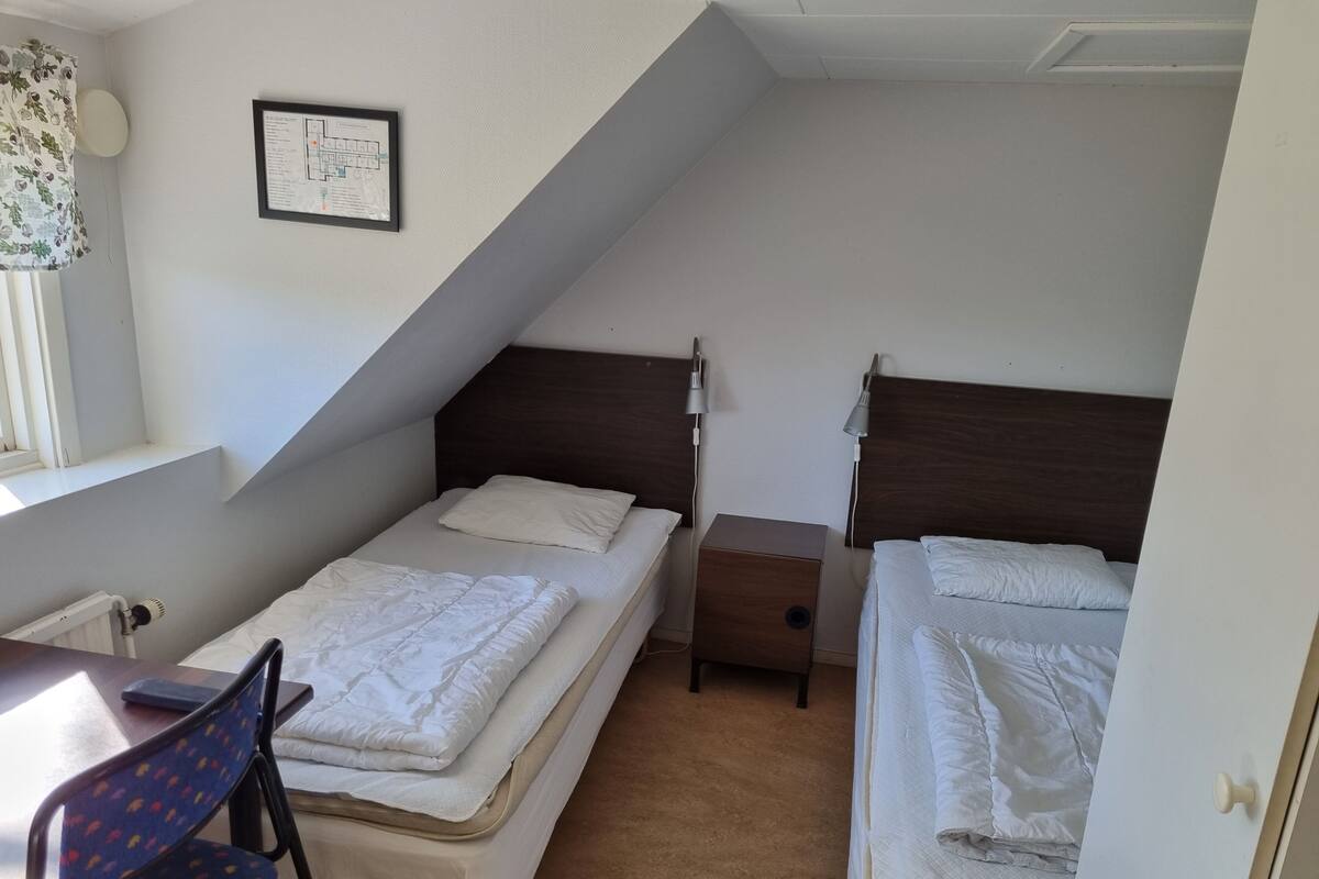 A small, well-lit room with two single beds, a desk, a chair, and a framed map on the wall.