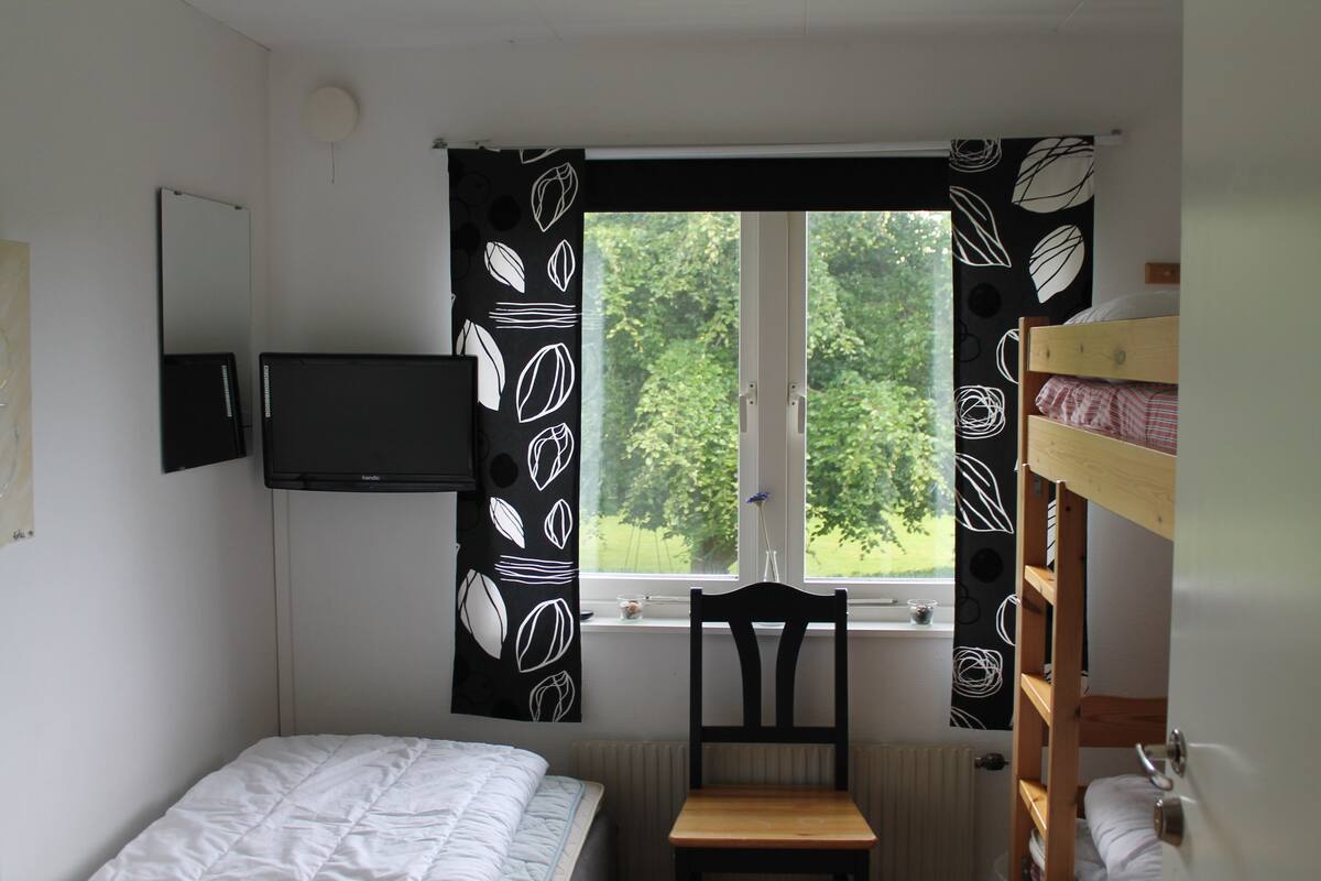 A bedroom with a bunk bed, a single bed, a chair, a TV mounted on the wall, and a window with curtains.