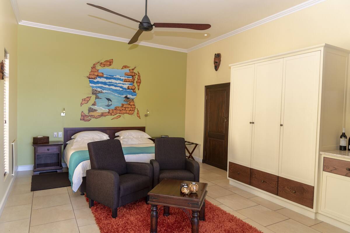 A bedroom with a bed, two chairs, a coffee table, a wardrobe, and a ceiling fan.