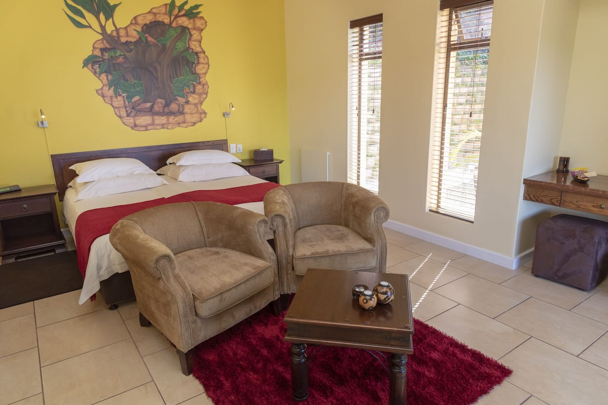 A hotel room with a bed, two armchairs, a small table with a red rug, and a wall mural.