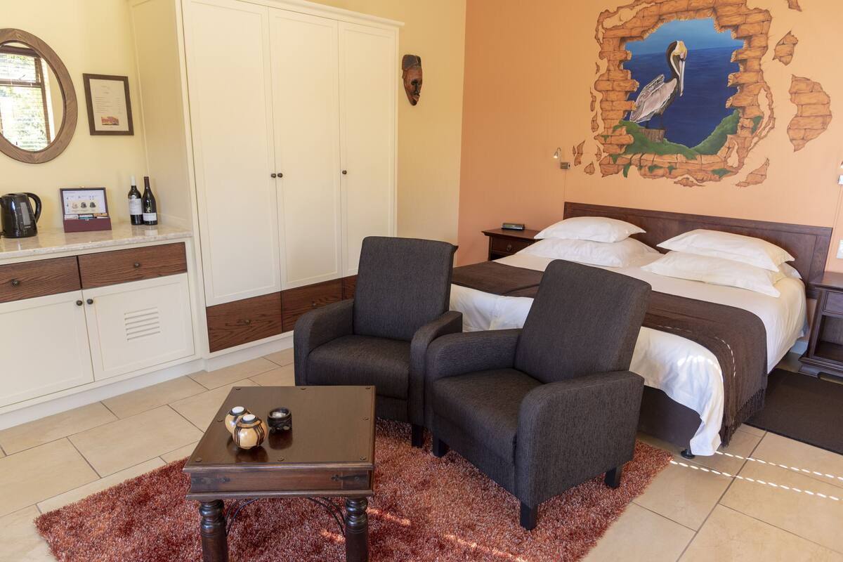 A bedroom with a bed, two armchairs, a wooden coffee table, a dresser, a mirror, and a wall mural.