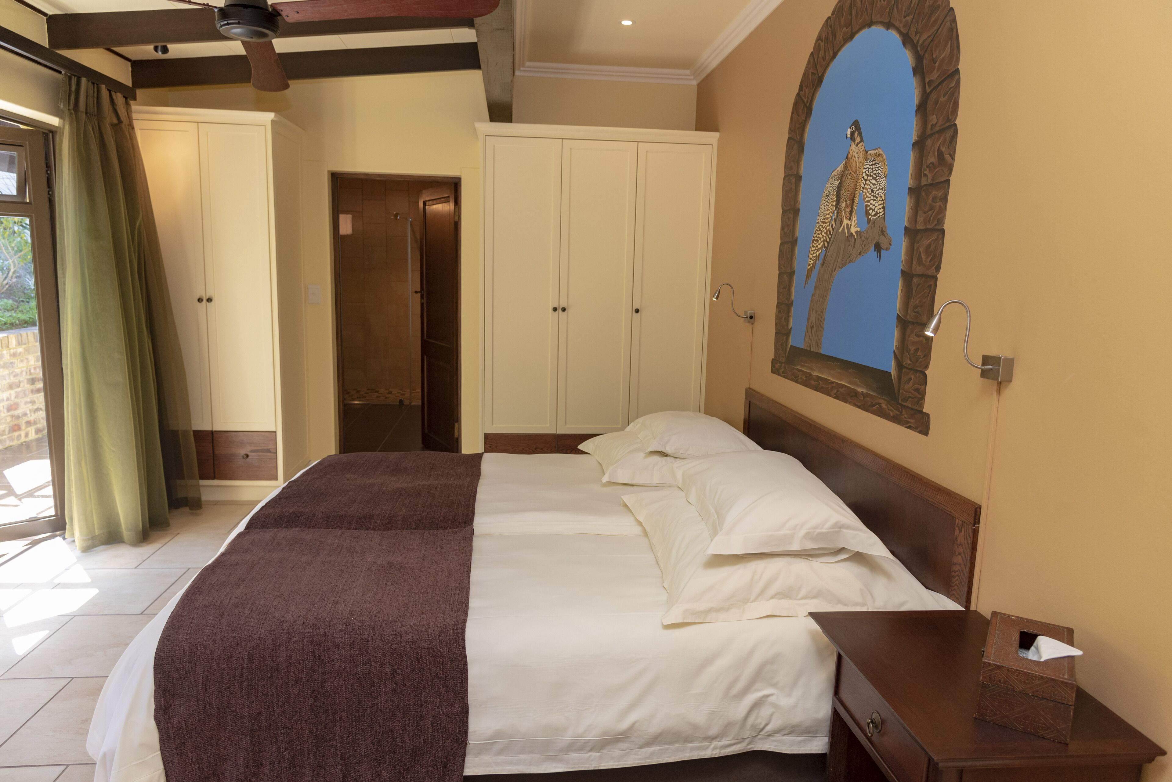 A bedroom with a large bed, a wooden nightstand, a mirror with a bird painting, and a door leading to another room.