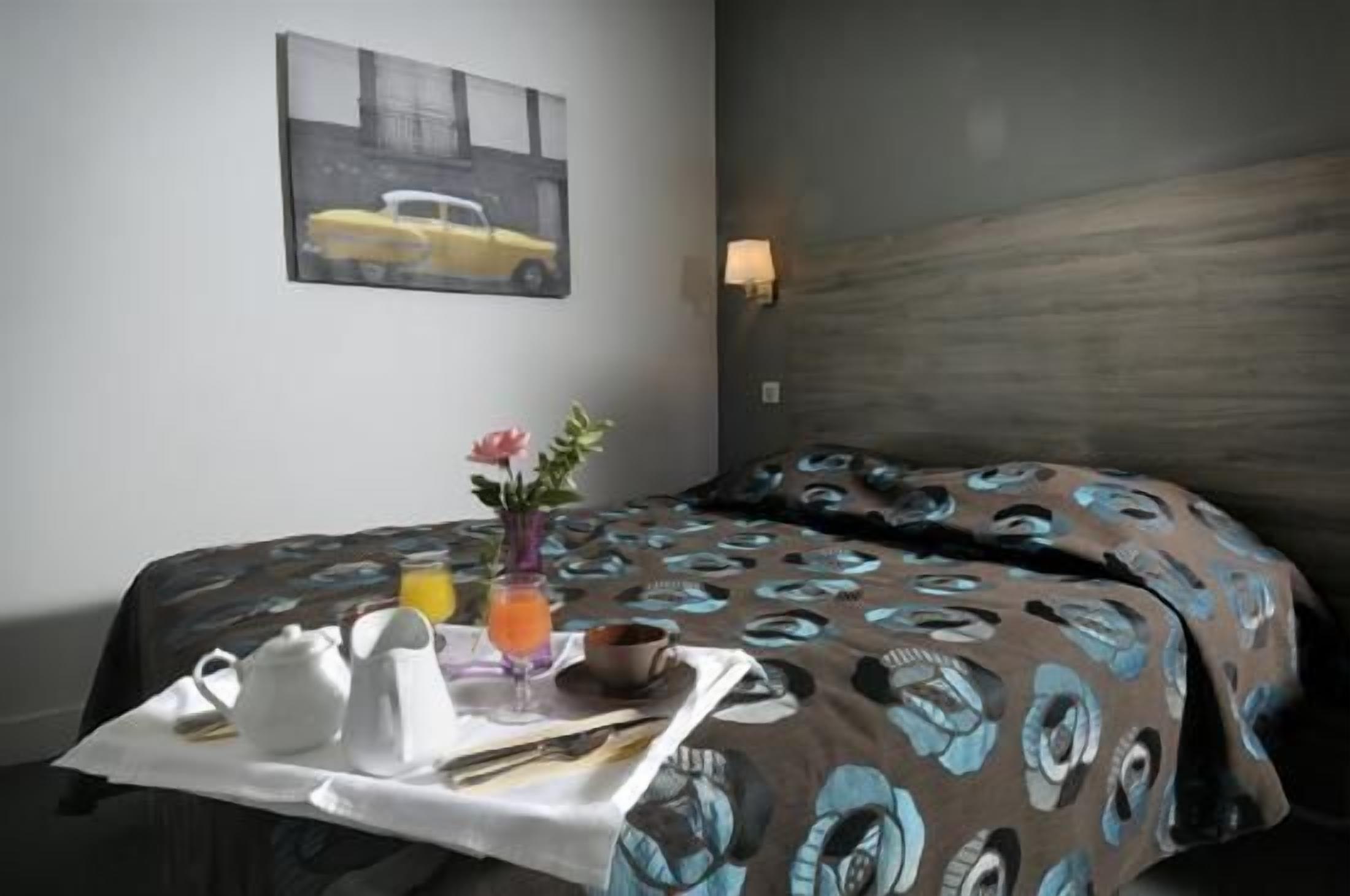 A hotel room with a bed, a table with breakfast items, a vase with flowers, and a painting on the wall.