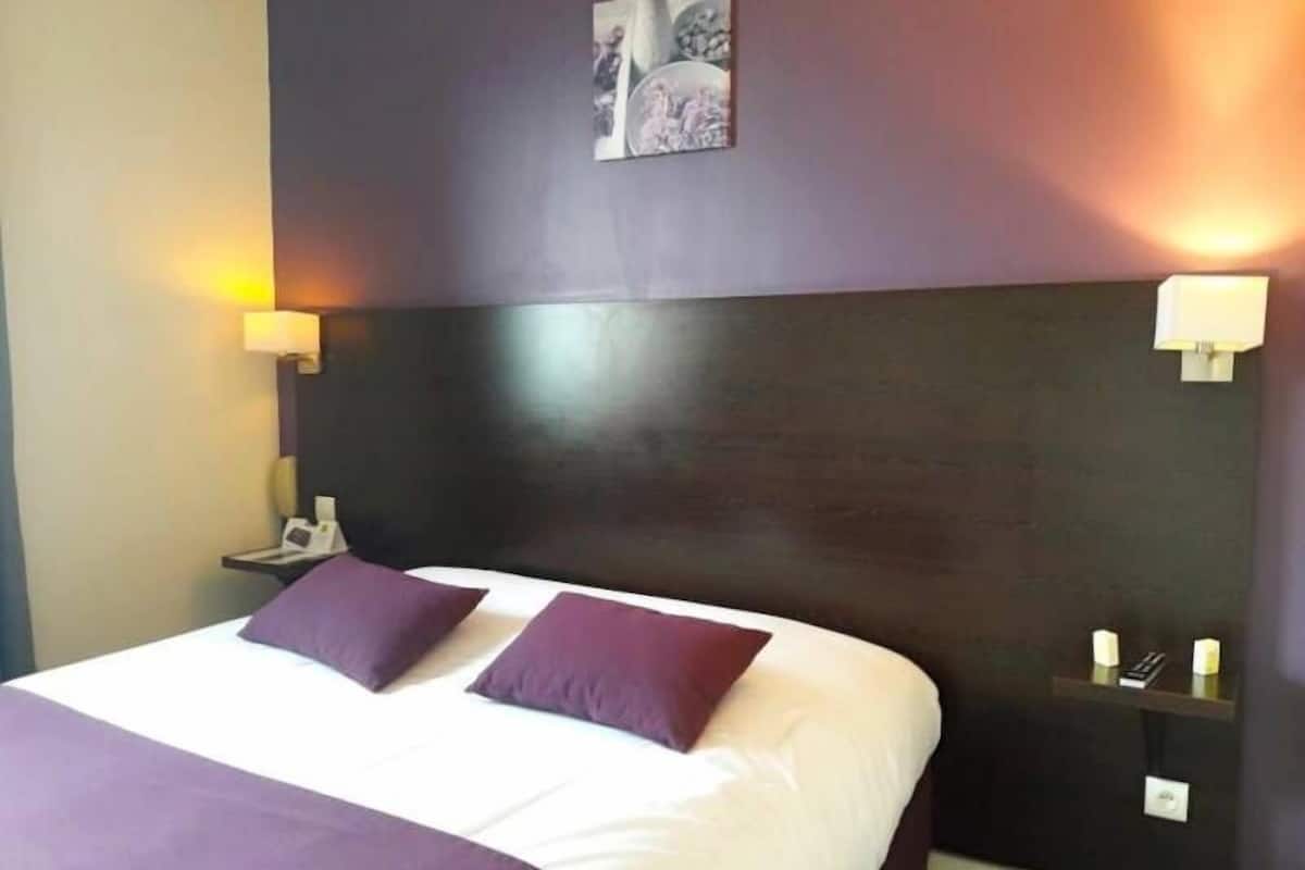 A hotel room with a large bed, two pillows, a nightstand, a wall-mounted lamp, and a small shelf with candles.