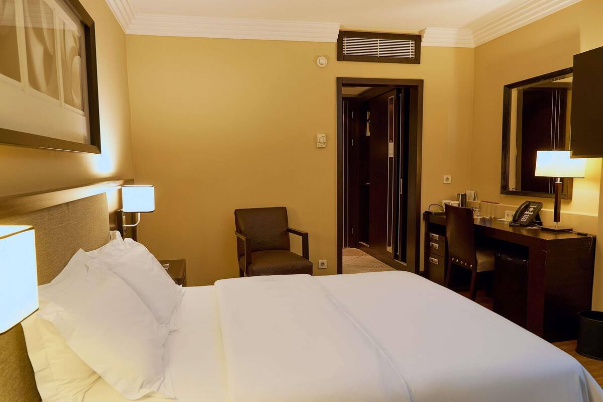 A hotel room with a bed, a desk, a chair, a mirror, and a door leading to another room.