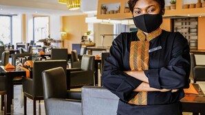 A person standing in a modern restaurant with high chairs and tables, pendant lighting, and a neutral color scheme. - Hotel Pullman Lubumbashi Grand Karavia (Lubumbashi)