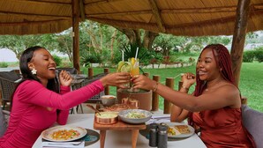 Two people toasting at a table with food and drinks, surrounded by a thatched roof and greenery. - Hotel Pullman Lubumbashi Grand Karavia (Lubumbashi)