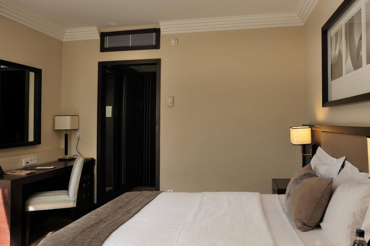 A hotel room with a bed, bedside table, lamp, desk, chair, and a door leading to another room.