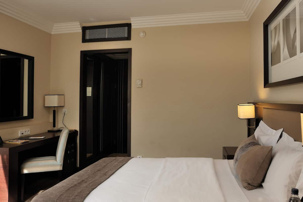 A hotel room with a bed, bedside table, lamp, desk, chair, and a door leading to another room.