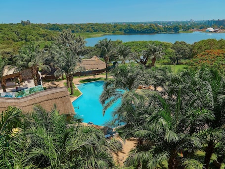 A resort with a swimming pool surrounded by palm trees and a view of a lake.. Hotel Pullman Lubumbashi Grand Karavia