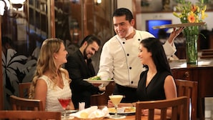 Une salle de restaurant avec des tables et des chaises en bois, un serveur qui apporte les plats et un vase rempli de fleurs sur la table.