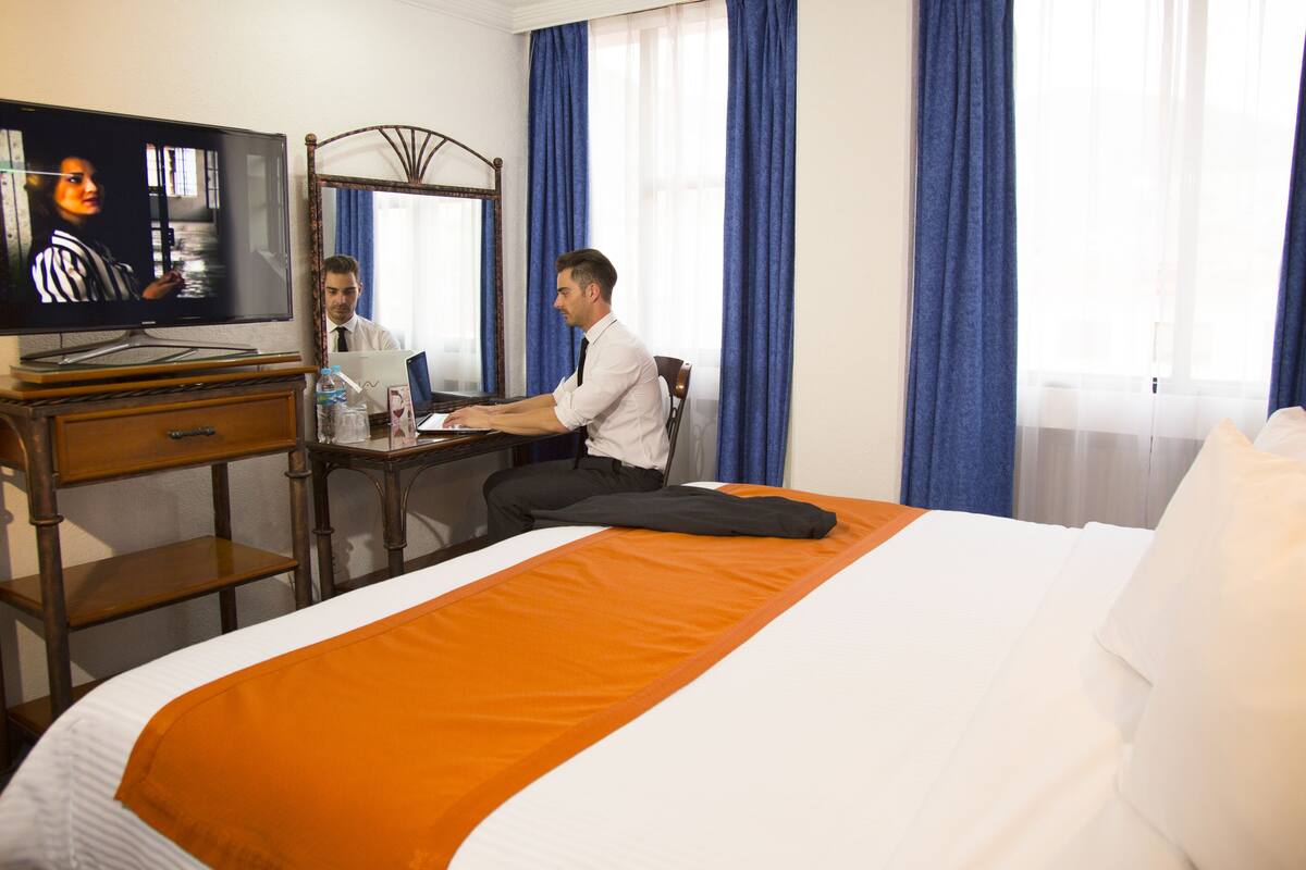 A hotel room with a bed, a desk with a laptop, a television, and a mirror.