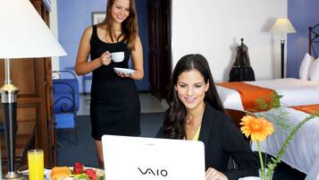 A hotel room with a bed, a desk with a laptop, a dining table with breakfast, and a lamp.