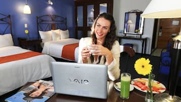 A hotel room with a bed, a desk with a laptop, a magazine, a glass of green drink, and a plate of fruit.
