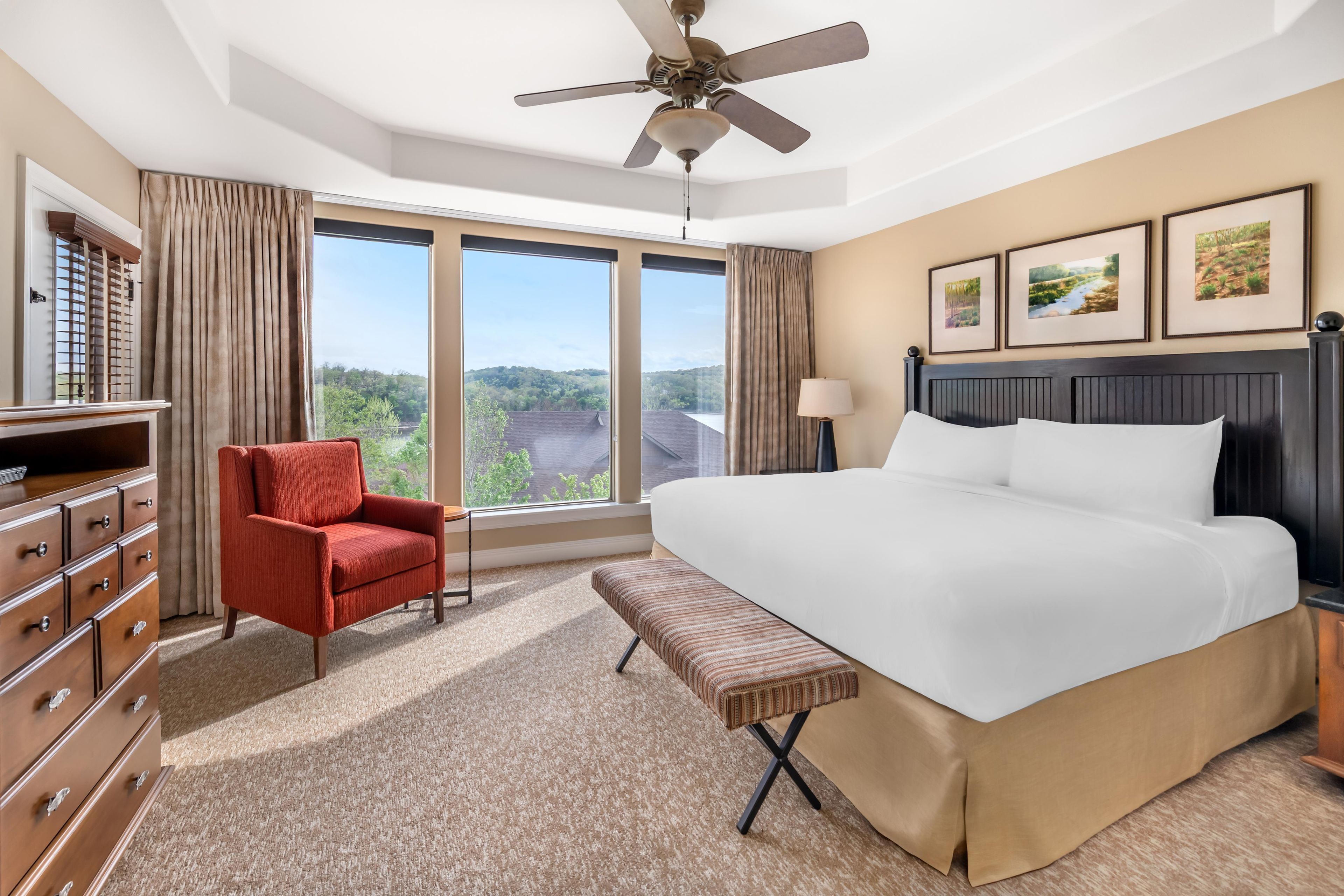 Habitación de hotel con una cama grande, un sillón rojo, un banco, un ventilador de techo y ventanas amplias con vistas al paisaje.