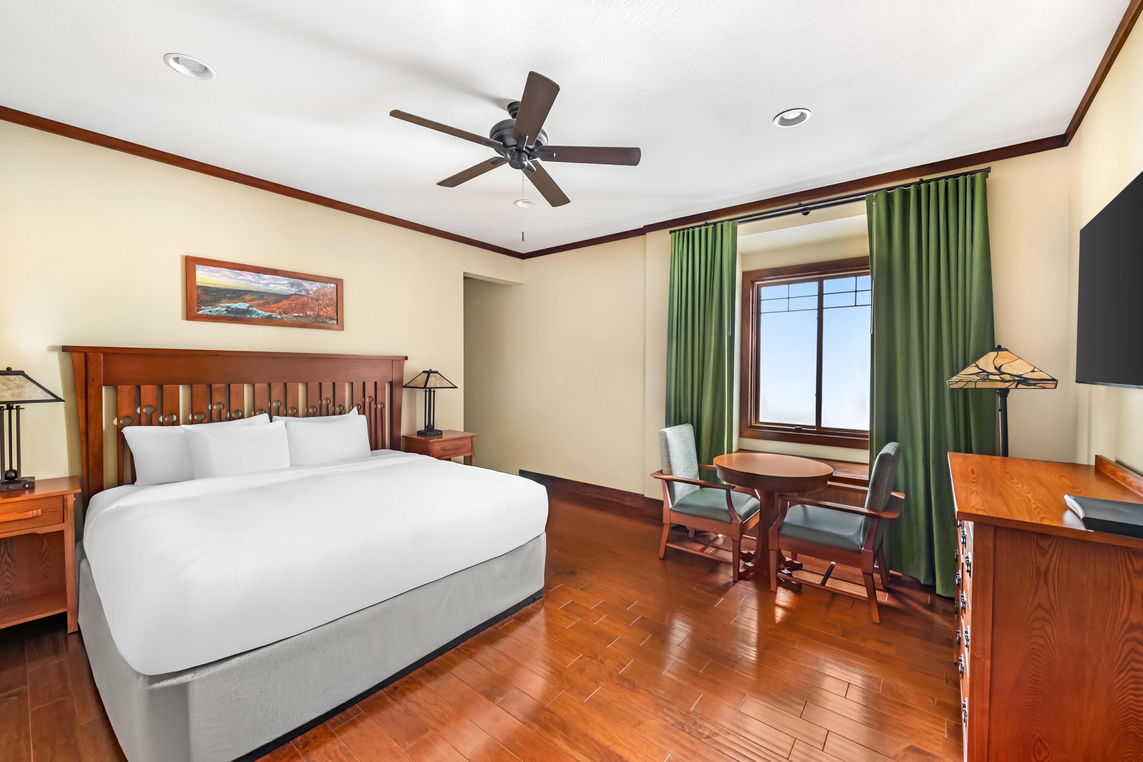 Quarto de hotel com cama, escrivaninha, cadeira, ventoinha no teto e um quadro na parede.