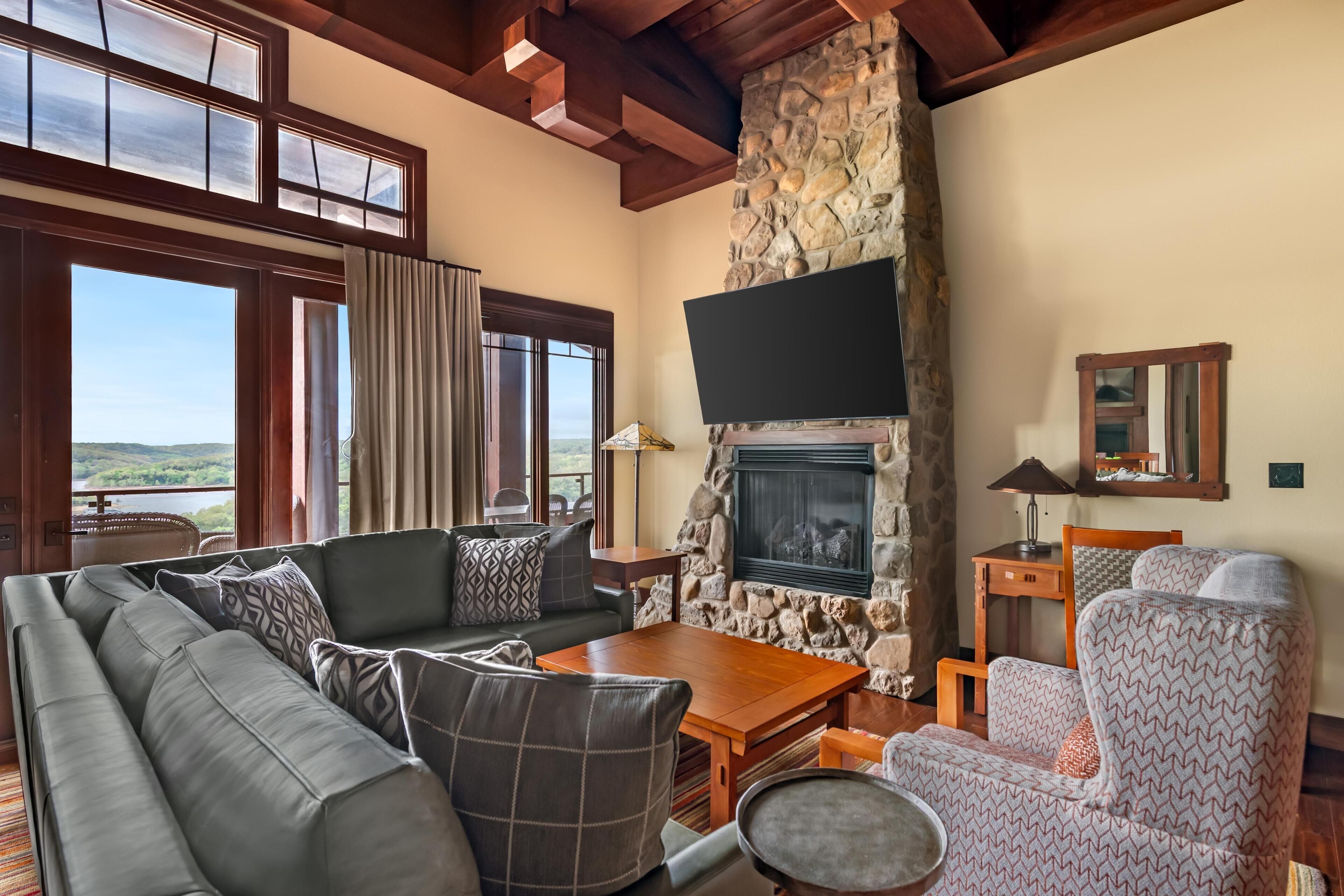 Un salón con chimenea de piedra, un ventanal con cortinas, un sofá, un sillón estampado y un televisor.