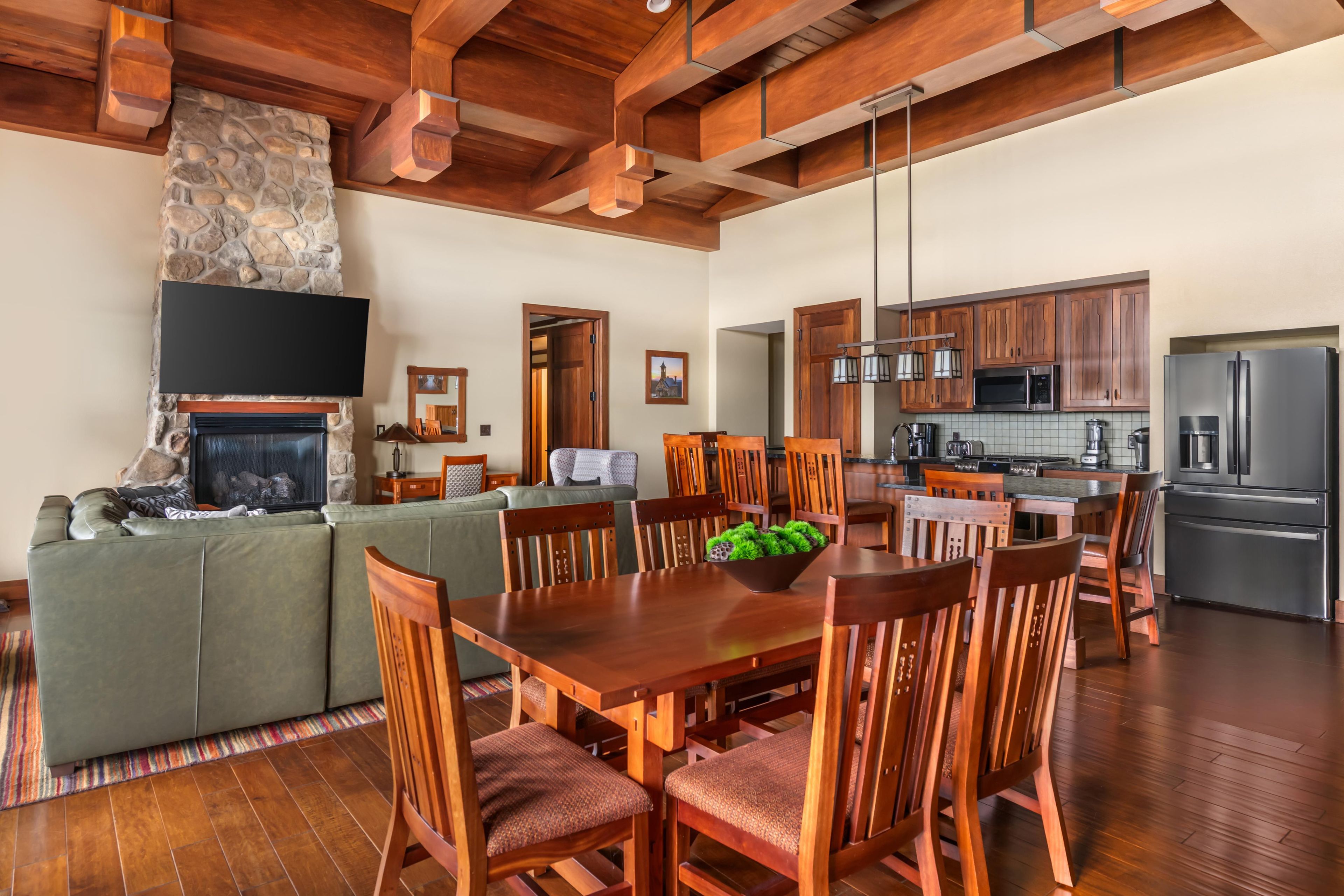 A spacious living area with a stone fireplace, wooden ceiling beams, a dining table with chairs, and a kitchen with modern appliances.