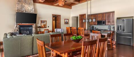 A spacious living area with a stone fireplace, wooden ceiling beams, a dining table with chairs, and a kitchen with modern appliances.