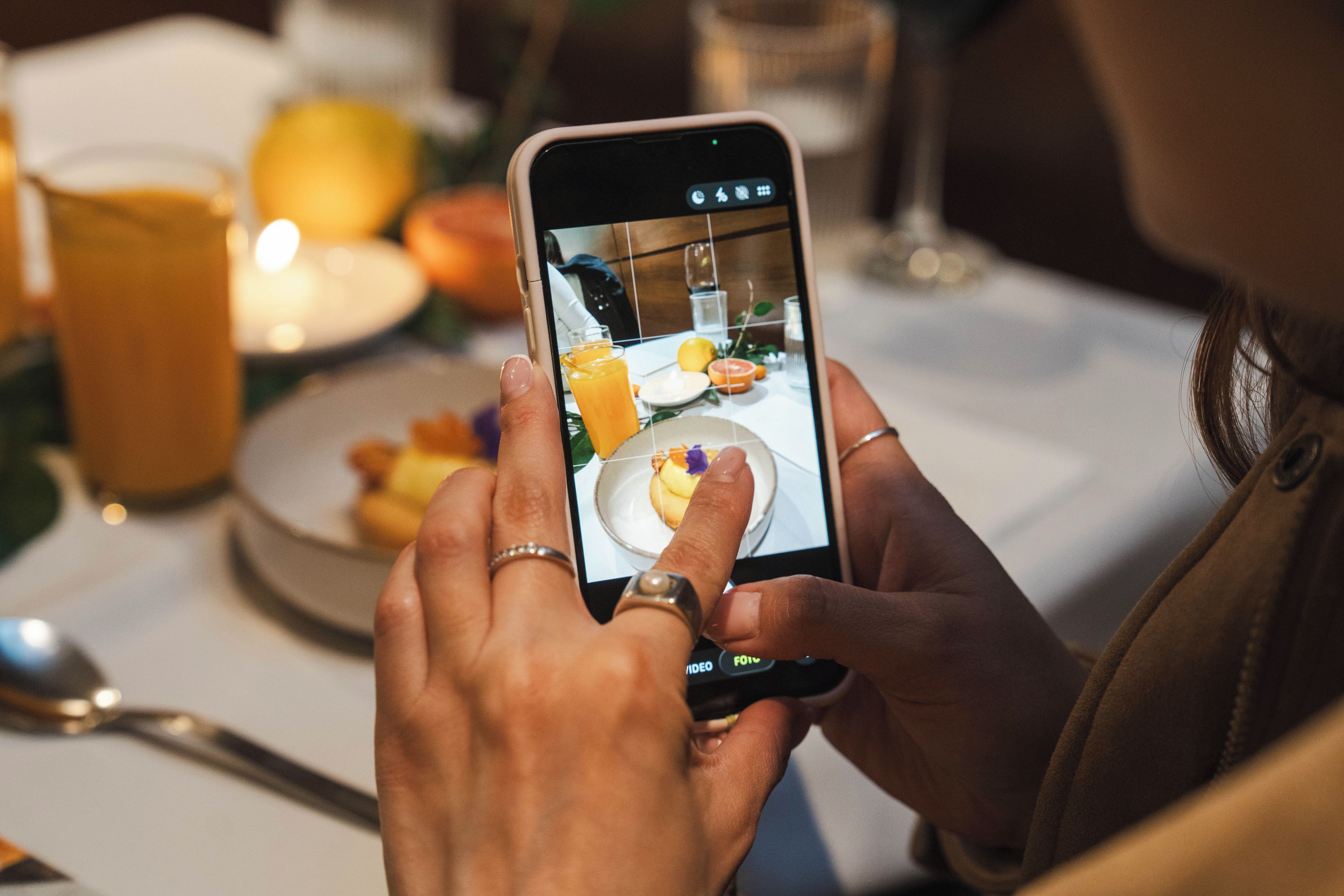 A person holding a smartphone, possibly taking a photo or video of a dining table with a glass of orange juice and a plate of food.